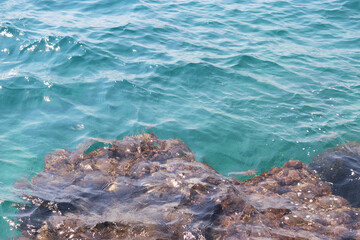 Turquoise sea waves break on rock on the seashore. Clear azure water on a stones covered with algae, natural background