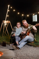 Young couple, caucasian woman and red haired bearded man, on a night picnic with a guitar and marshmello have a good time