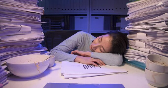 Exhausted overloaded asian business woman falling asleep on desk covered with stack of paperwork at home. Sleepy tired alone girl dozing off at the table while working hard at late night. Dolly shot.