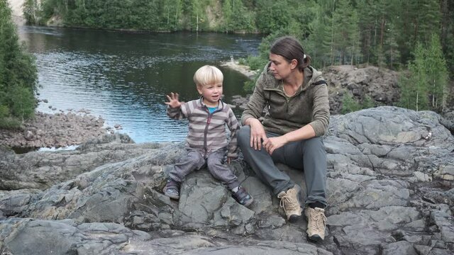 Mother and son hiking travellers sitting on cliff near mountain river and talking. Active family outdoors.