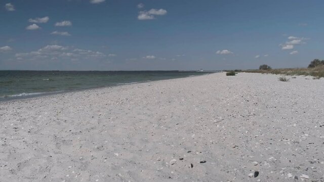 Sandy beach with shells near sea by sunny clear day. No peopele. Small sea waves hit sandy shore. Skyline with blue sky and white clouds extreme wide shot. Slow motion