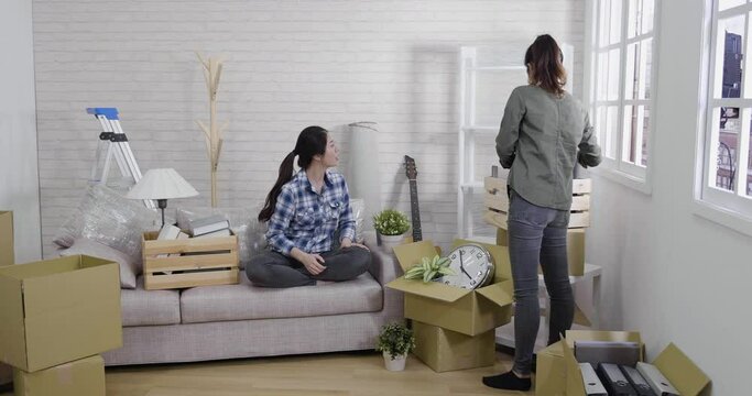 two asian japanese women friends removing stuff from moving box at new house living room. female roommates talking unpacking carton container cardboards with belongings in bright cozy apartment.