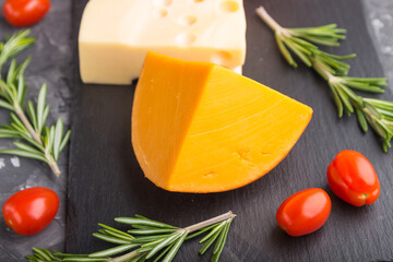 Various types of cheese with rosemary and tomatoes on black slate board on a black concrete background. Side view, close up.