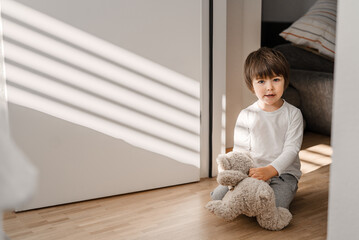 Cute little happy child playing with soft teddy bear toy sitting on wooden floor in room at home at sunny morning. Light and shadows.