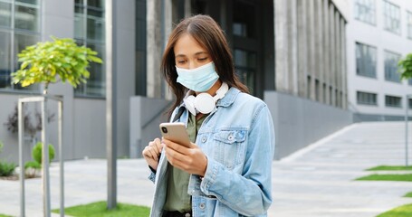 Pretty young Caucasian stylish girl student in medical mask and with backpack texting message on mobile phone. Beautiful female tapping and scrolling on smartphone. Pandemic concept. Outdoors.