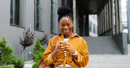 African American joyful young stylish woman tapping or scrolling on smartphone and standing at city...