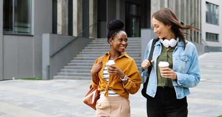 Mixed-races young pretty females best friends talking cheerfully and walking with cups of coffee to-go and at city street. Multi ethnic stylish happy women students strolling outside with hot drinks