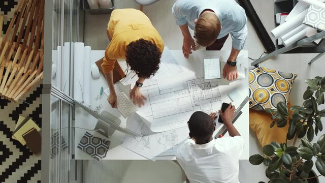 Top down shot of group of multiethnic male and female architects unrolling paper on office table, discussing construction plan and putting marks while working in team on project - Powered by Adobe