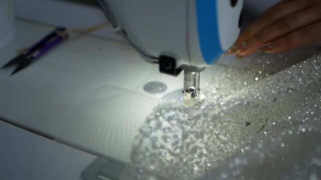 A woman with a beautiful manicure sews a wedding dress on a sewing machine. A textile worker puts white shiny fabric into a sewing machine. Garment factory for the manufacture of women's clothing.