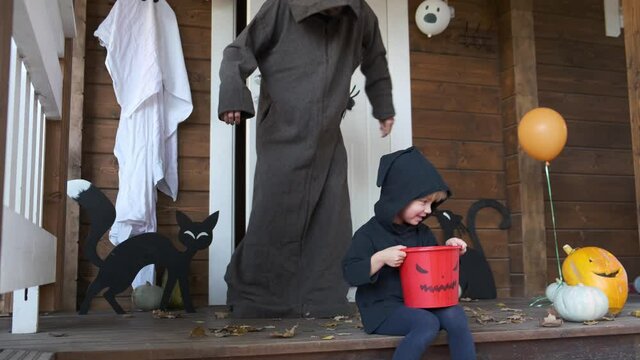 Halloween vibes. Cute child sitting near house door and looking at how much candy he collected in bucket while trick or treat, father in wizard costume suddenly grabs kid and scares him.