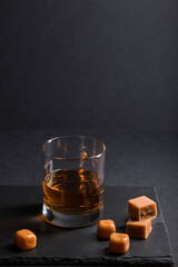 Glass of whiskey with caramel candies on a black stone slate board on black background. Side view,...