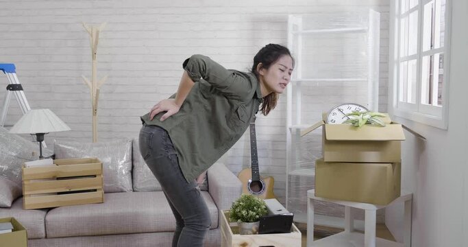 young asian woman suffering back ache moving boxes in new house living room. girl hurt painful waist while lifting large carton cardboard relocation. injured bad posture wrong spine pain sick bone