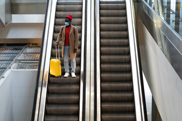 Top view of Black man with yellow luggage standing on escalator at airport terminal wearing face protective mask during virus epidemic, covid-19 pandemic. Second wave coronavirus, new normal concept