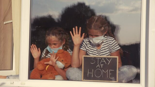 stay at home coronavirus. kids in medical mask in home with a toy teddy bear. kids holding a sign with the inscription stay at home. quarantine sitting by the window. coronavirus covid 19 concept