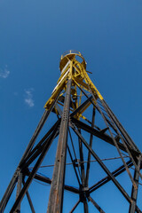 a photograph of an old somewhat rusty lighthouse painted black and yellow with a blue sky background and vertically cropped