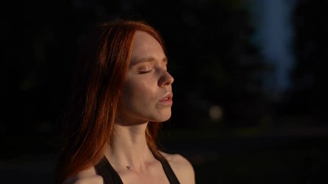 Close-up face with closed eyes of attractive meditative young woman with emotion of serene and calm, meditating outside in city park background of bright sun lights in morning at sunrise.