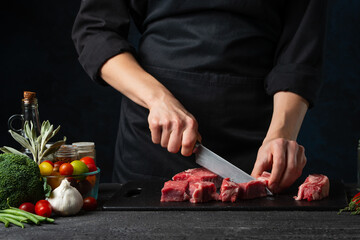 The professional chef cutting with knife fresh beef meat on the black board on dark blue...