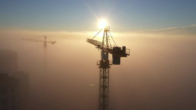 Construction site at dawn. Tower cranes over fog on the background of the morning sun. Drone view