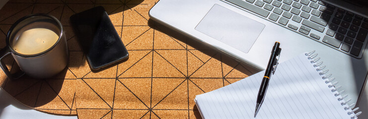 Ángulo elevado de un escritorio moderno con una plancha de corcho de figuras geométricas bajo la luz del sol de la mañana. Oficina en el hogar con laptop, teléfono móvil, anotador y una taza de café.