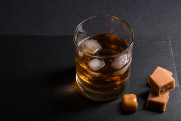 Glass of whiskey with ice and caramel candies on a black stone slate board on black background. Side view, low key, copy space.