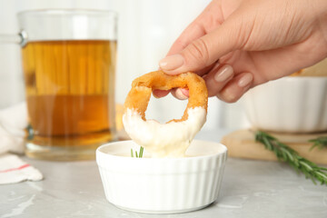 Woman dipping crunchy fried onion ring in sauce at grey marble table, closeup