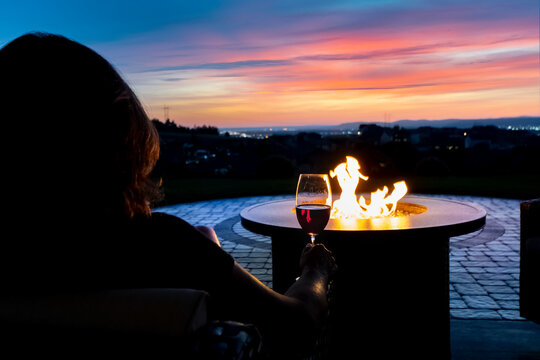 A woman relaxes with a glass of wine at sunset by a fire pit on the patio of a luxury home overlooking the Spokane valley and city in Spokane, Wa USA