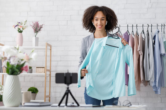 Advertising new collection in blog of modern fasion and working from home. Smiling african american lady with wireless headphones showing blue shirt and looking at webcam