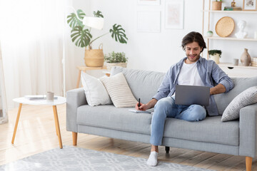 Fototapeta premium Young man with laptop taking notes, home interior