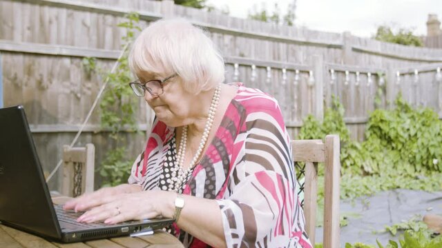 Elderly Woman Typing On A Black Laptop Slowly Thinking And Working From Home Outside Garden