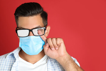 Man wiping foggy glasses caused by wearing medical mask on red background. Space for text