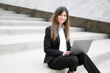 Beautiful businesswoman in suit, using a laptop sitting on stairs outdoors. High quality photo.
