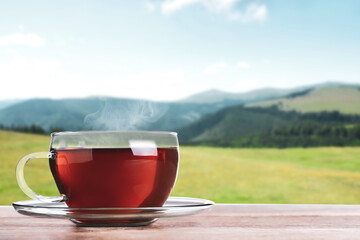 Glass cup of fresh hot tea on wooden table against blurred mountain landscape. Space for text
