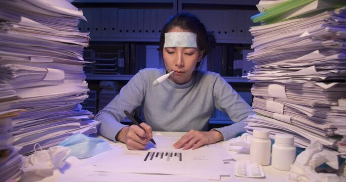 Sick asian business woman is sitting at desk covered with stack of paperwork and have cooling gel sheet on head. Alone girl checking temperature with thermometer while working hard at late night.