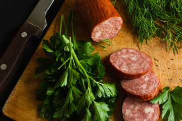 pieces of smoked sausage, parsley and dill on a cutting Board. Slicing on the light wooden cutting board with kitchen knife against the black background