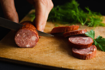 slicing smoked sausage and with a kitchen knife on a cutting board made of light wood with parsley...