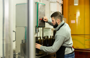 Naklejka na ściany i meble Skilled guy working on conveyor for bottling olive oil in glass bottles in small family factory