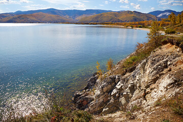 Autumn landscape with beautiful yellowed trees on the shore of lake Baikal on a sunny day. Calm clear water, pebble beach.