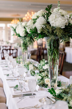 The head table at a wedding reception with tall glass trumpet vases filled with white flower and green arrangement for a classic wedding design. 