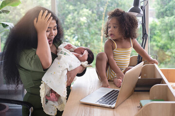 Dark Skinned mother cares for her newborn baby and stressed by the information on her Laptop, newborn baby (16 days old) sleeps in her mother's arms, little girl sits on a table, selective focus