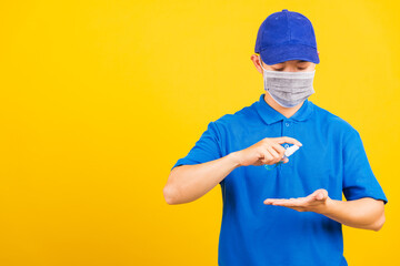 Asian young handsome delivery man wearing face mask protective germ virus and mask alcohol sanitizing sprays to hand, studio shot isolated on yellow background, medical outbreak coronavirus COVID-19
