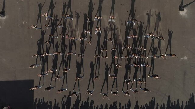 Top down aerial view of a large group of young people dancing outdoors on asphalted area. Shadow dance. A crowd of spectators nearby. Flash mob. 
