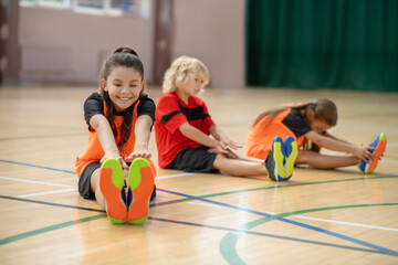 Three kids exercising in the gym and stretching froward