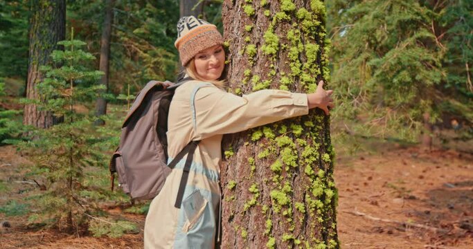 Nature lover is hugging cinematic tree bark covered with scenic green moss on sunny autumn day. Pretty female traveler exploring fall nature. Environmental activist is wrapping hands around tree trunk