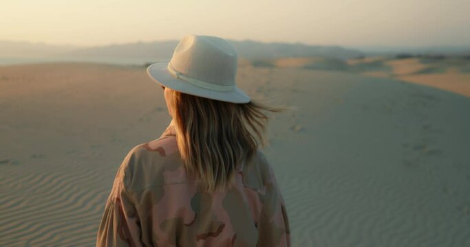 Slow motion woman enjoying golden sunset over ocean. Beautiful female walking by the sand dunes and looking on the sun in clear sky. Lady in stylish clothes on outdoor adventure. Golden Hour footage