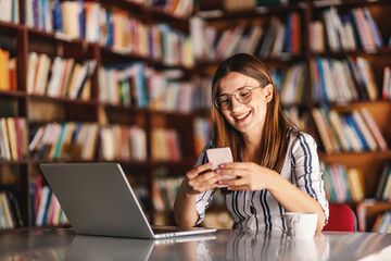 Young smiling brunette with eyeglasses sitting in library and using smart phone. There is a laptop...