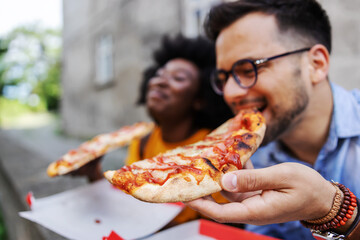 Closeup of young multicultural hipster couple sitting outdoors and eating pizza. Selective focus on...