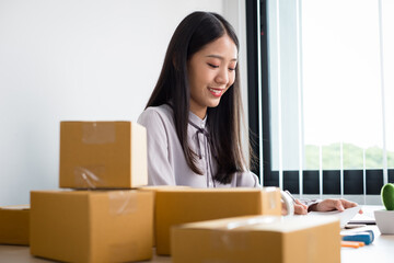 Fototapeta na wymiar Young Asian woman business owner with many parcel boxes on the table happy online sales job, use your laptop, get an order from customers, take notes, and make arrangements for delivery by post