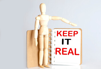 Wooden man shows with a hand to white notepad with text keep it real