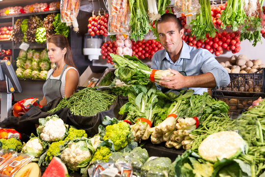 Female shop assistant weighing vegetables on scales while male worker laying out vegetables at vegetable shop