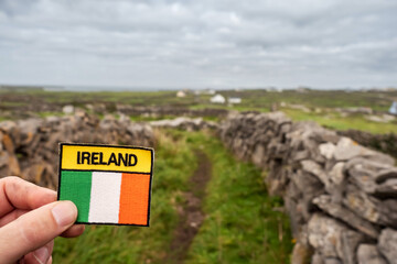 Tourist holding badge with sign Ireland and Irish National flag in focus, Blurred landscape in the background, Traditional stone fences. Inis mor Aran Islands, county Galway, Ireland. © mark_gusev
